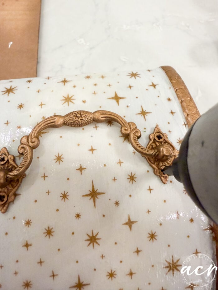 A hand uses a glue gun to attach a gold handle to a white box decorated with gold stars on a marble surface, showcasing a simple Flipping Smalls DIY upgrade.