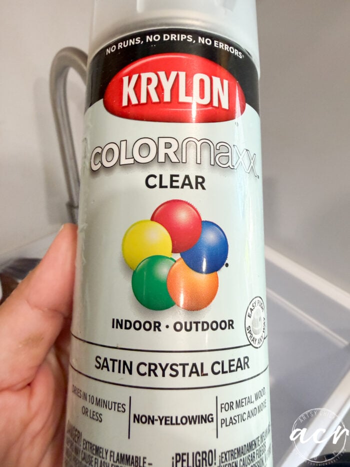 Hand holding a can of Krylon COLORmaxx Clear spray paint, labeled Satin Crystal Clear. The can, perfect for Flipping Smalls projects, features colorful spheres and is suitable for indoor and outdoor use.