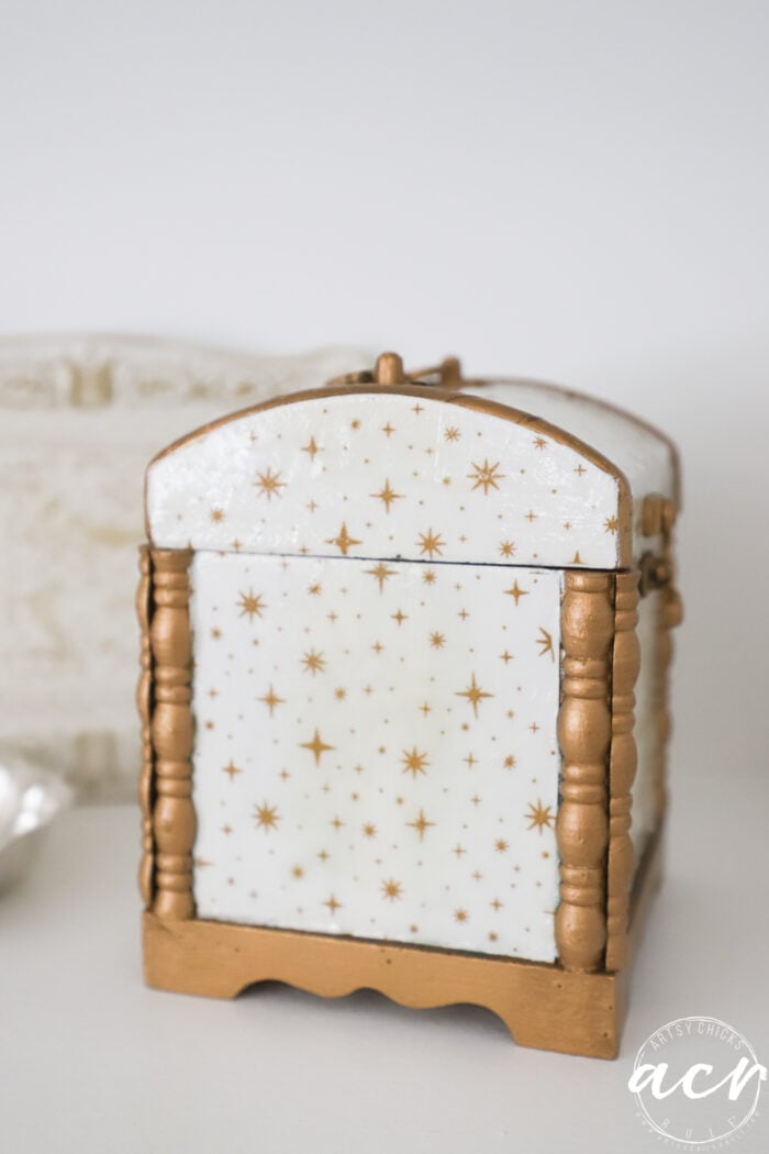 A small decorative box from Flipping Smalls with a white background and gold star patterns, featuring gold-painted trim and legs, sits on a white surface.