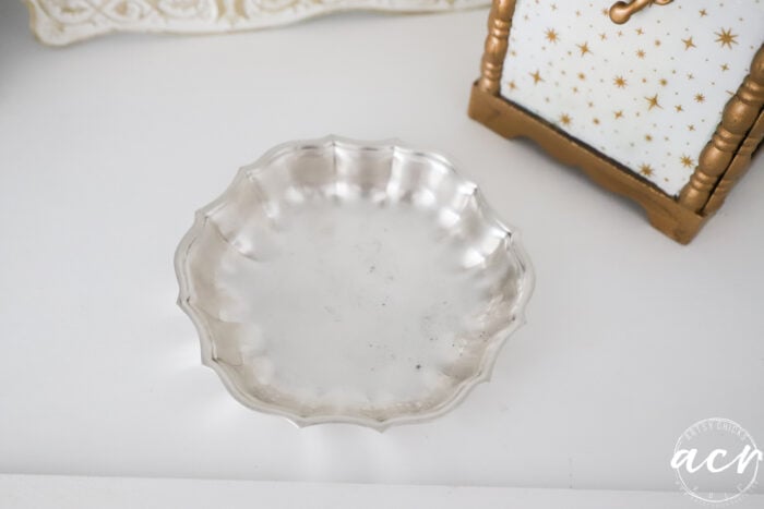 A small, scalloped-edge silver dish sits on a white surface near a decorative gold and white item with star patterns, making it a charming addition to any Flipping Smalls collection.