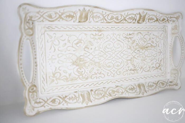 A rectangular, ornate white tray with intricate, weathered beige designs and handles on each side. The tray has a vintage look, perfect for adding a touch of Flipping Smalls charm to your décor. The bottom right corner has a white circular logo with acy written in it.