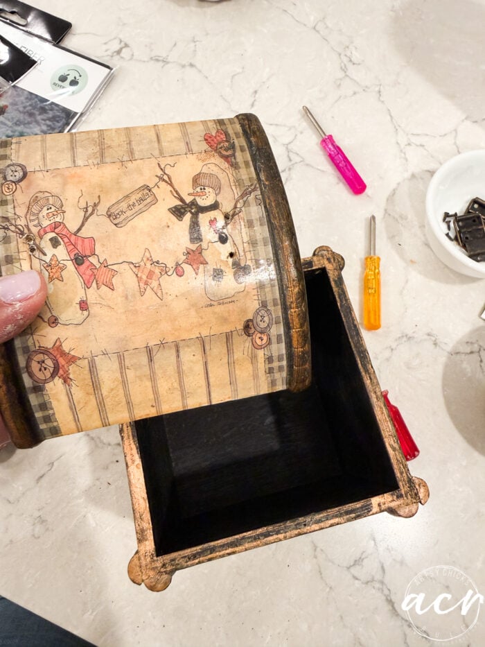 A hand holds a painted wooden box lid decorated with snowmen, stars, and stripes above an open, empty box on a marble surface with tools and hardware nearby—a charming moment from Flipping Smalls #4.