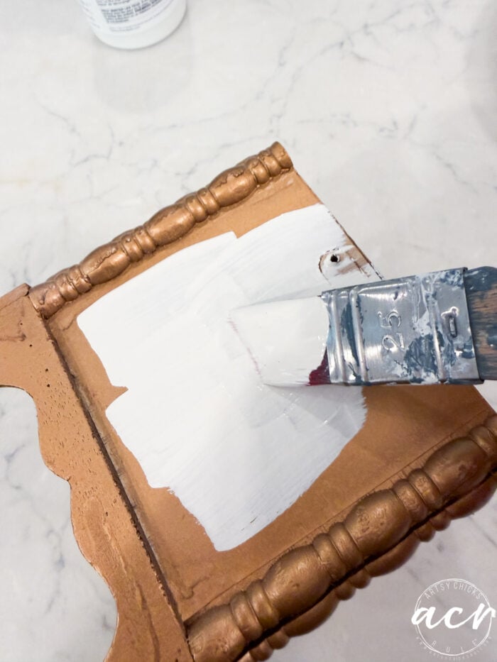 A paintbrush applies white paint to a small, ornate wooden surface with a gold finish, on a marble countertop—a perfect transformation for Flipping Smalls #4 collectibles.