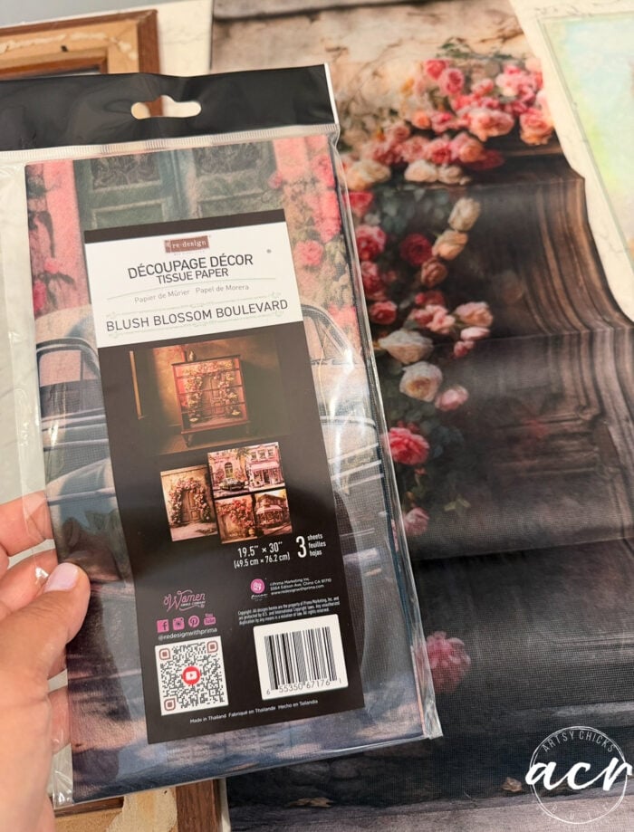 A hand holds an updated package of tissue paper labeled Découpage Décor – Blush Blossom Boulevard. Floral-patterned tissue paper and a floral art piece, along with framed prints, are visible in the background on a table.