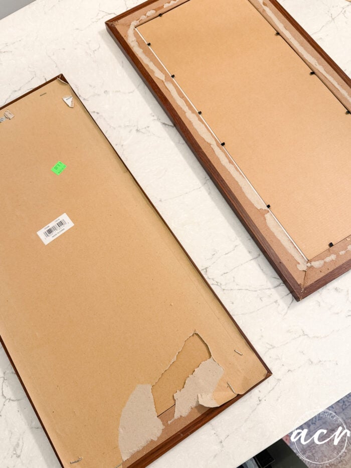 Two rectangular picture frames with brown backing lie on a white marble surface; the backing of one frame is torn, exposing the inside. A green price sticker and a barcode sticker are visible, suggesting these framed prints have recently been updated or unwrapped from their original tissue paper packaging.