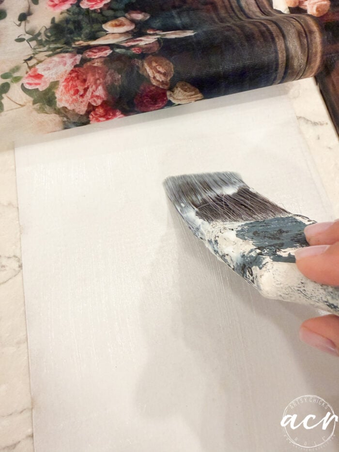 A hand uses a paintbrush to apply white glue on a flat surface, preparing it for a floral sheet with pink and burgundy flowers, partially rolled back—an ideal technique for creating decorative tissue paper projects or custom framed prints.