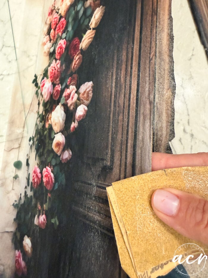 A hand holding sandpaper smooths the edge of a wooden board featuring an image of pink and red roses climbing up a brown door, as if preparing it for display alongside elegant framed prints.