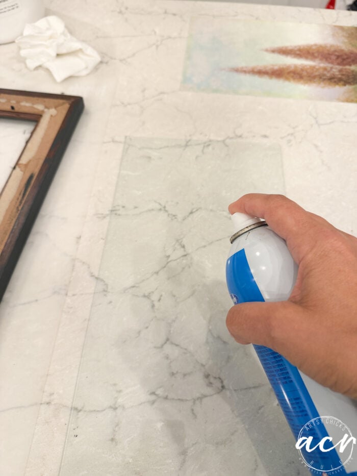 A hand sprays a clear substance from a blue and white can onto a pane of glass, which is placed on a marble-patterned surface. A wooden frame, tissue paper, and artwork for framed prints are nearby, giving the scene an updated look.