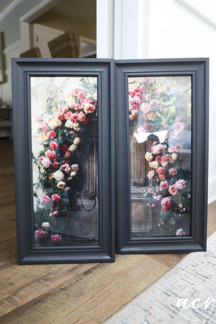 Two black-framed prints featuring bouquets of pink and red roses arranged on a rustic wooden door, leaning against a white wall on a wooden floor, bring an updated touch to any space.