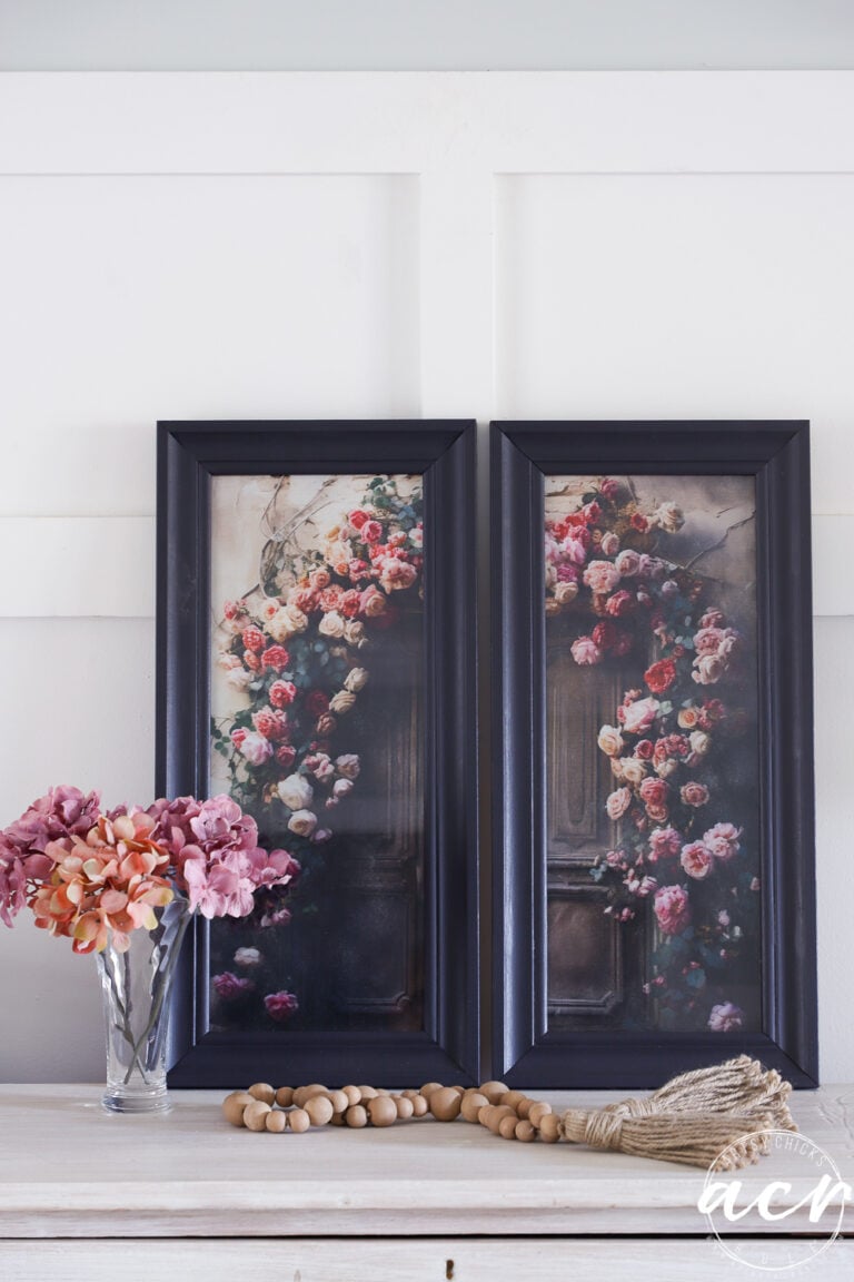 Two updated framed prints featuring floral designs lean against a white wall above a white table, which holds a glass vase with pink and mauve flowers, wooden beads, and a decorative tassel.