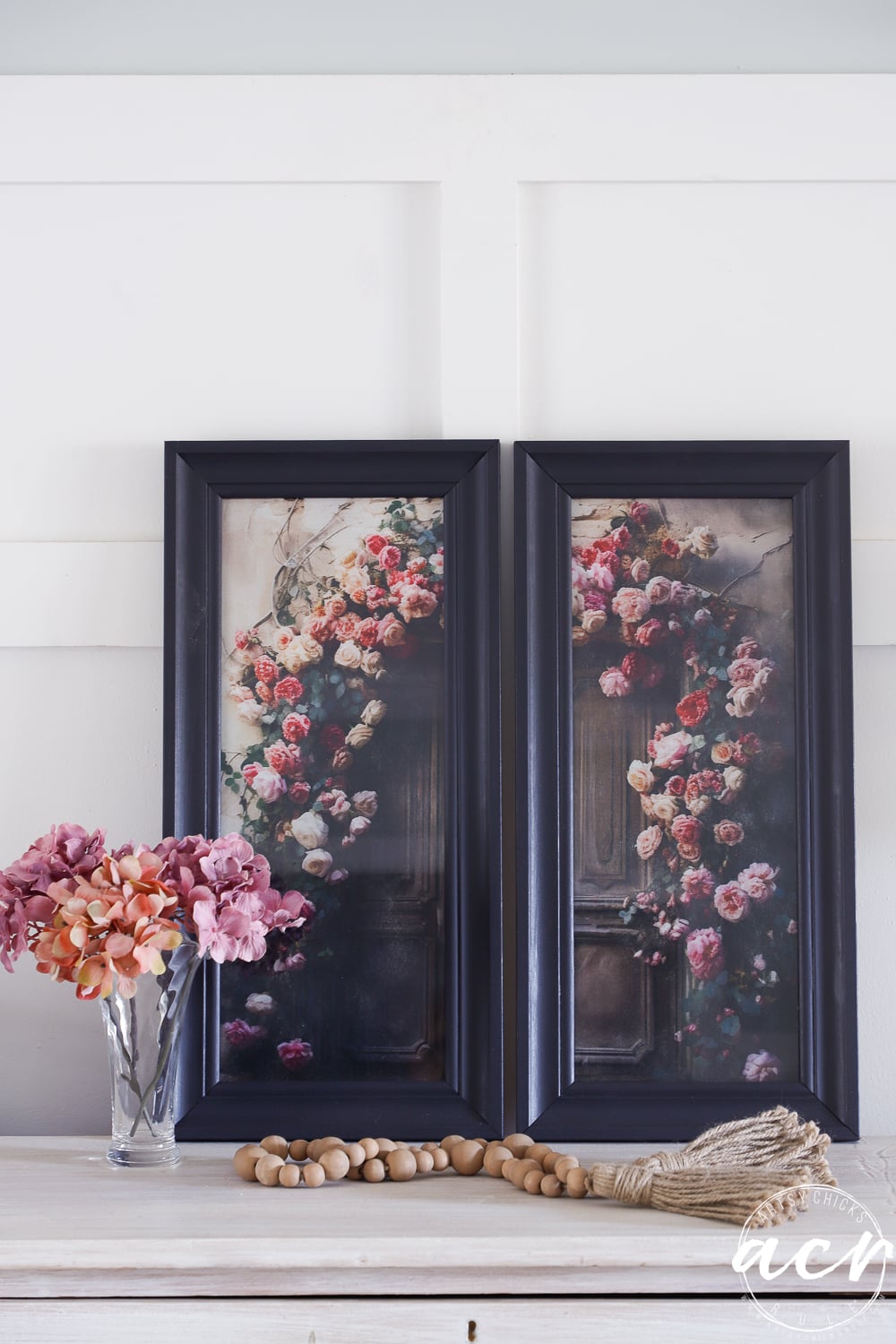 Two updated framed prints featuring floral designs lean against a white wall above a white table, which holds a glass vase with pink and mauve flowers, wooden beads, and a decorative tassel.