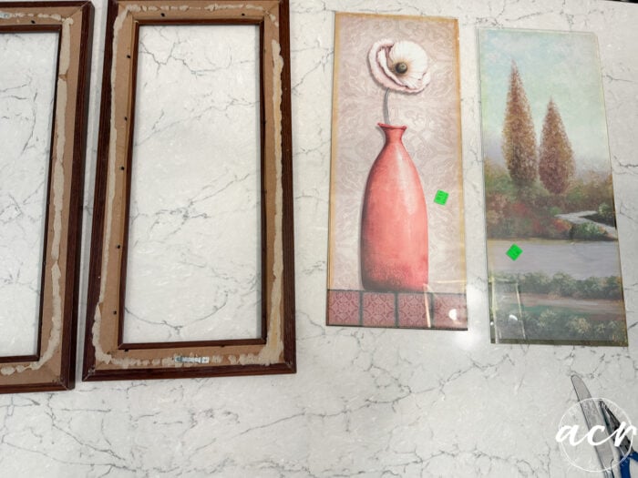 Two empty wooden frames and two decorative wall art pieces—including updated framed prints, one featuring a red vase with a white flower and another depicting trees by a lake—are all arranged on a white marble surface.