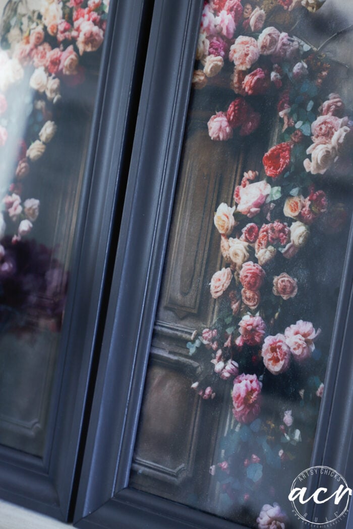A close-up of dark-framed glass cabinet doors featuring a floral design with cascading pink and red roses and green leaves, reminiscent of delicate tissue paper art or elegant framed prints.