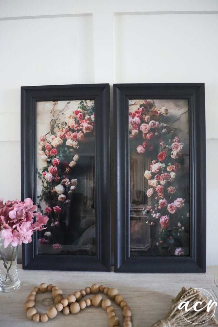 Two black-framed floral prints with pink and white roses are propped side by side on a light wood table. A glass vase with pink flowers, a beaded garland, and delicate accents crafted from tissue paper add an updated touch to the tabletop arrangement.