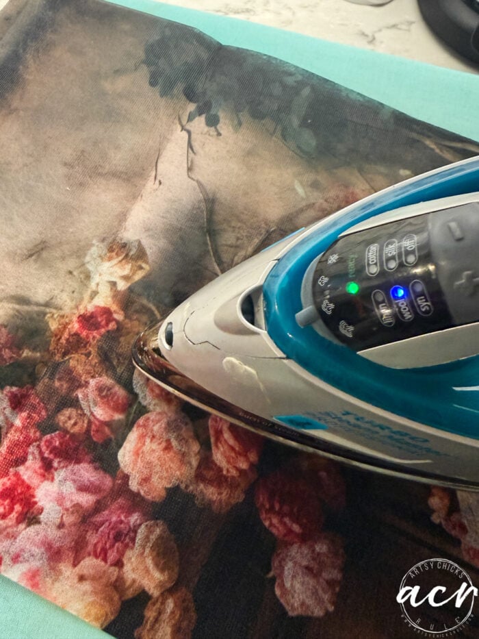 A close-up of an iron pressing a floral-patterned fabric on a light blue surface, with the artist’s signature logo visible in the bottom right corner. This updated piece is also available as framed prints for a refined display.