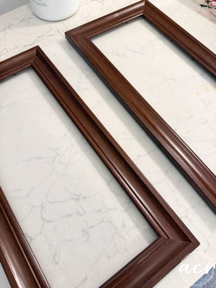 Two empty rectangular wooden picture frames with a dark finish are placed on a light-colored marble surface, ready to display updated framed prints of your choice.