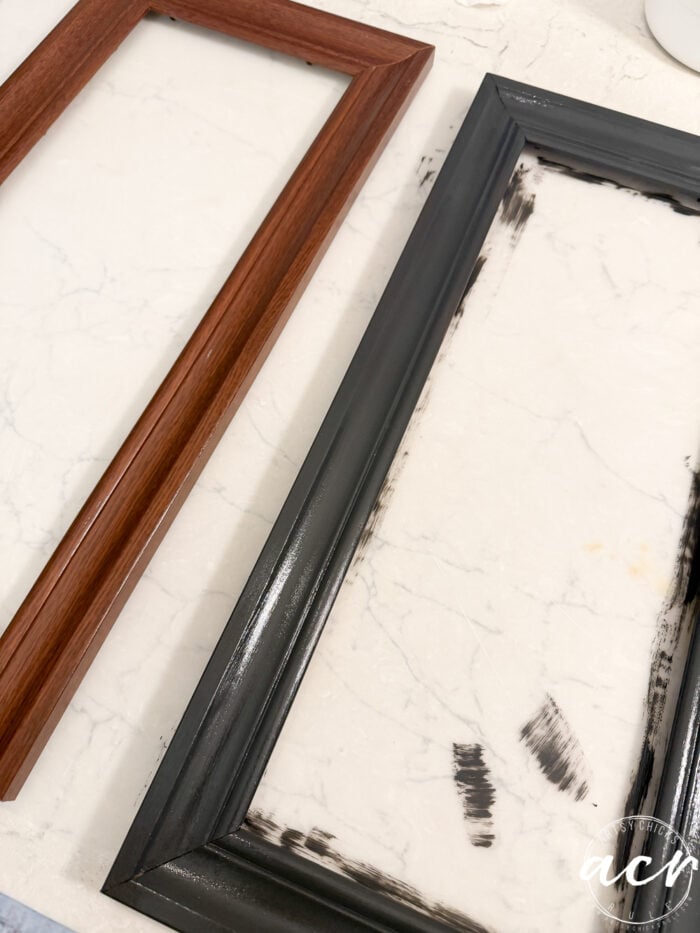 Two empty picture frames lie on a white marble surface; one frame is brown wood, the other, ideal for displaying updated framed prints, is partially painted black with some brush strokes visible on the marble.
