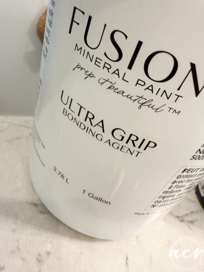 A close-up of a white container labeled FUSION MINERAL PAINT ULTRA GRIP BONDING AGENT, 3.78 L, 1 Gallon, sitting on a light-colored countertop—an essential product for updated décor projects like tissue paper crafts or refreshing framed prints.