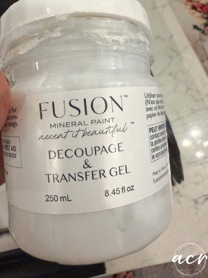 A hand holds a clear plastic jar labeled Fusion Mineral Paint Decoupage & Transfer Gel, 250 mL, 8.45 fl oz. The jar contains a white gel, perfect for use with tissue paper or updating framed prints. A marble surface and part of a brush are visible in the background.