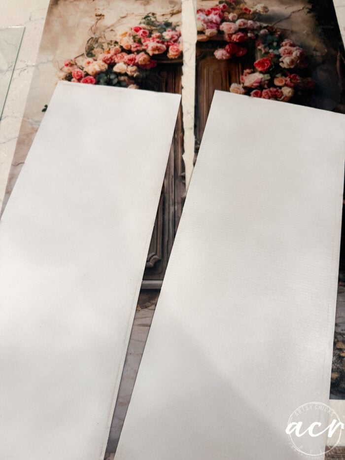 Two white rectangular boards, resembling tissue paper in their delicate appearance, are placed on top of a decorative image featuring a wooden door with an arch of pink and white flowers. The boards partially cover the door and floral design, evoking the layered look of framed prints overlapping a beautiful backdrop.