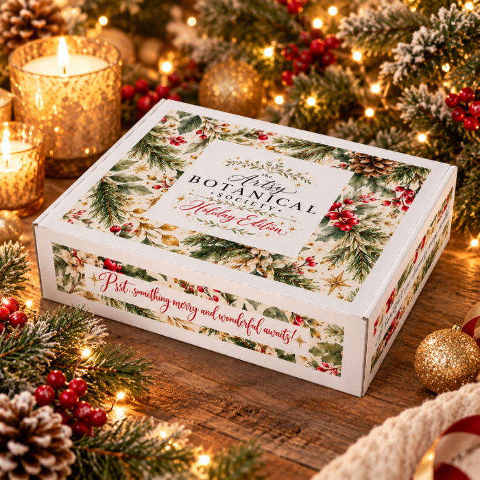 A rectangular Holiday Edition box from The Artsy Botanical Society, surrounded by Christmas ornaments, candles, and evergreen branches that highlight seasonal Christmas favorites.