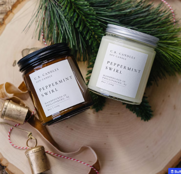 Two C.B. Candles Peppermint Swirl soy candles rest on a wood slice with pine needles and small bells, perfect for your Christmas favorites.