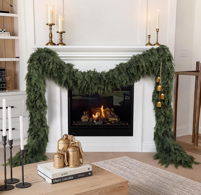 Green cedar garland draped over a white fireplace mantel, decorated with hanging gold bells and pillar candles as one of our Christmas favorites.