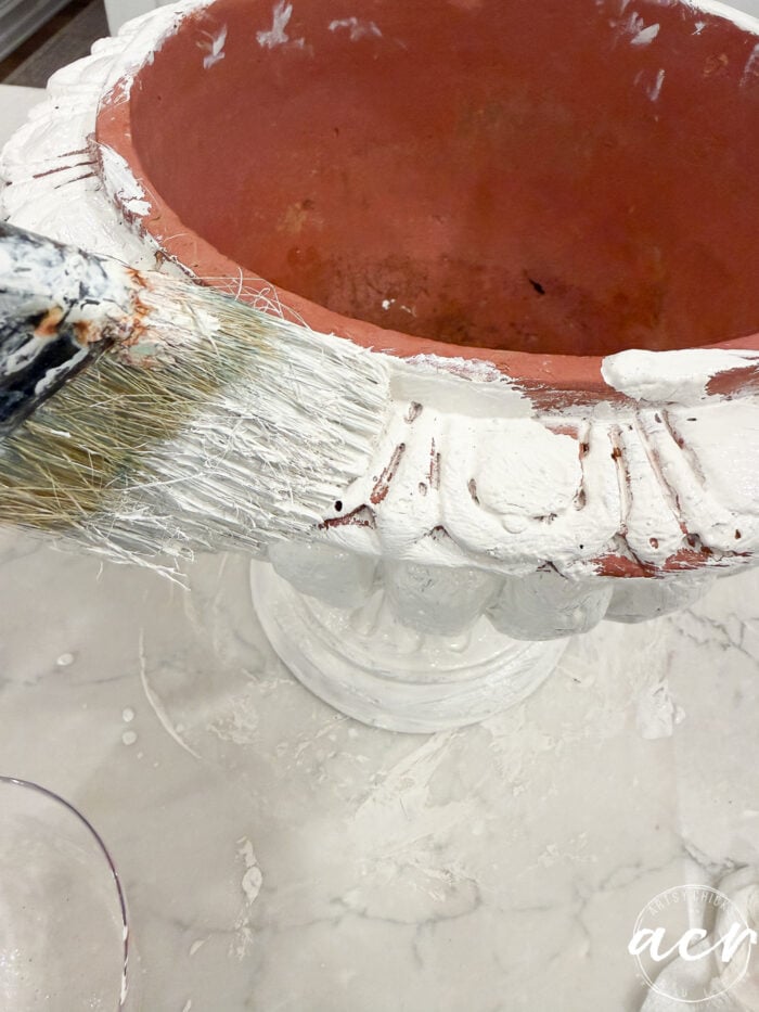 A paintbrush applying white paint to the decorative rim of a terracotta urn during a DIY concrete planter tutorial.