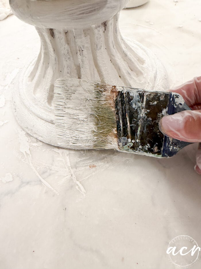 A gloved hand uses a brush to apply white paint to the base of a decorative pedestal for a DIY concrete planter project.