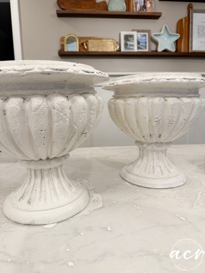 Two white pedestal urns with a distressed, faux concrete finish sit on a marble surface.