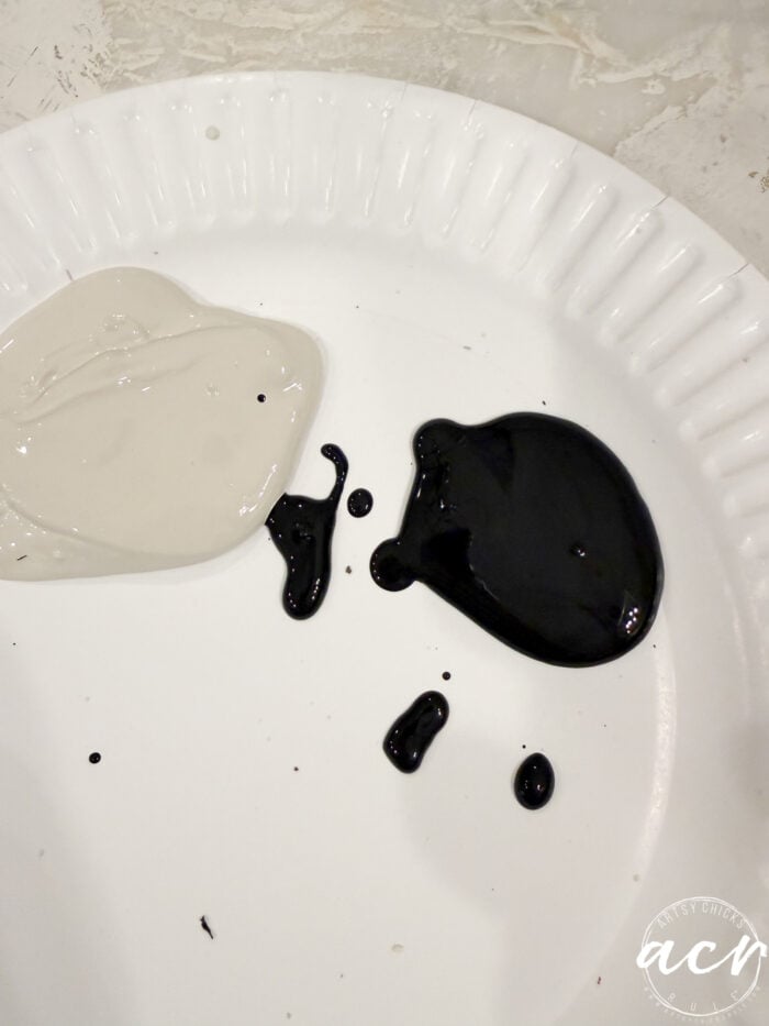A dollop of beige paint and a dollop of black paint on a white paper plate, ready to be mixed for a faux concrete project.
