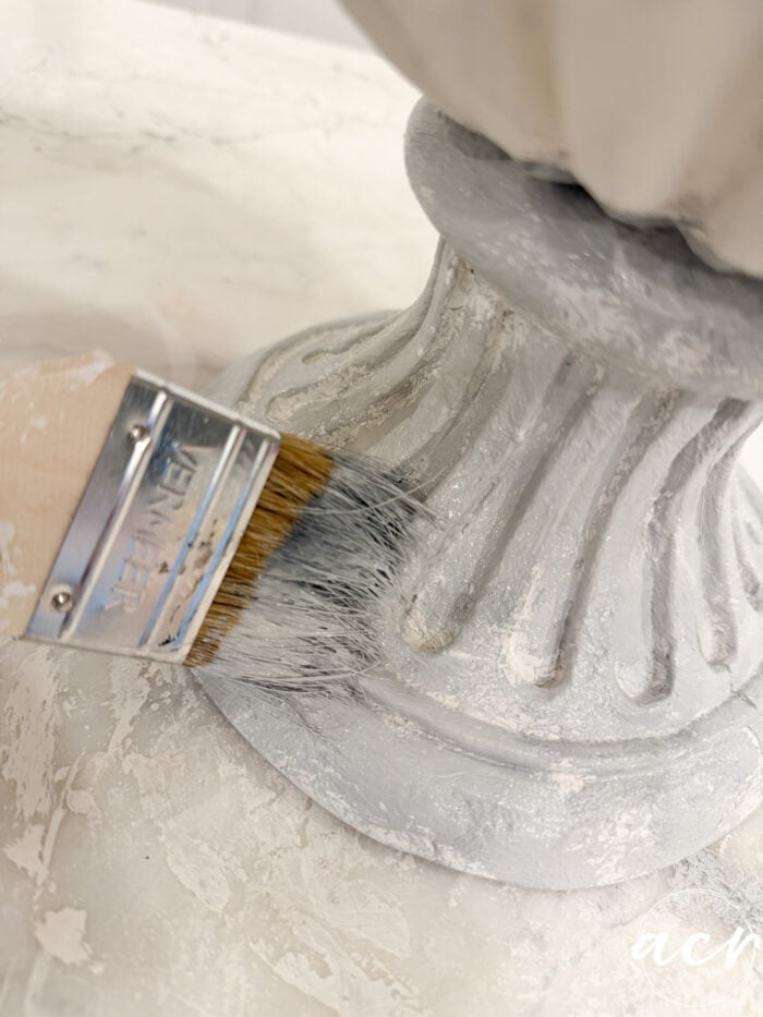 A paintbrush applying light grey paint to the base of a fluted decorative urn for DIY concrete planters.