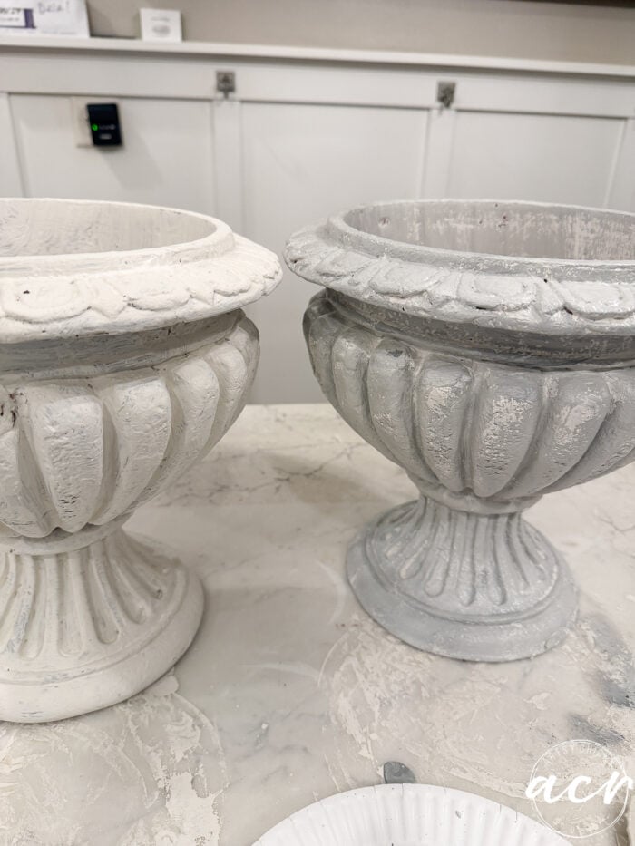 Two decorative pedestal urns sitting on a counter, one painted matte white and the other finished with a DIY concrete planter effect in weathered gray.
