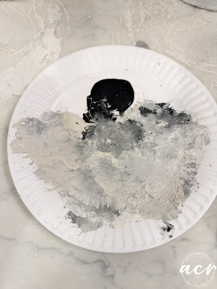 A paper plate used as a palette, containing swirls of black, white, and various shades of gray acrylic paint for DIY concrete planters.
