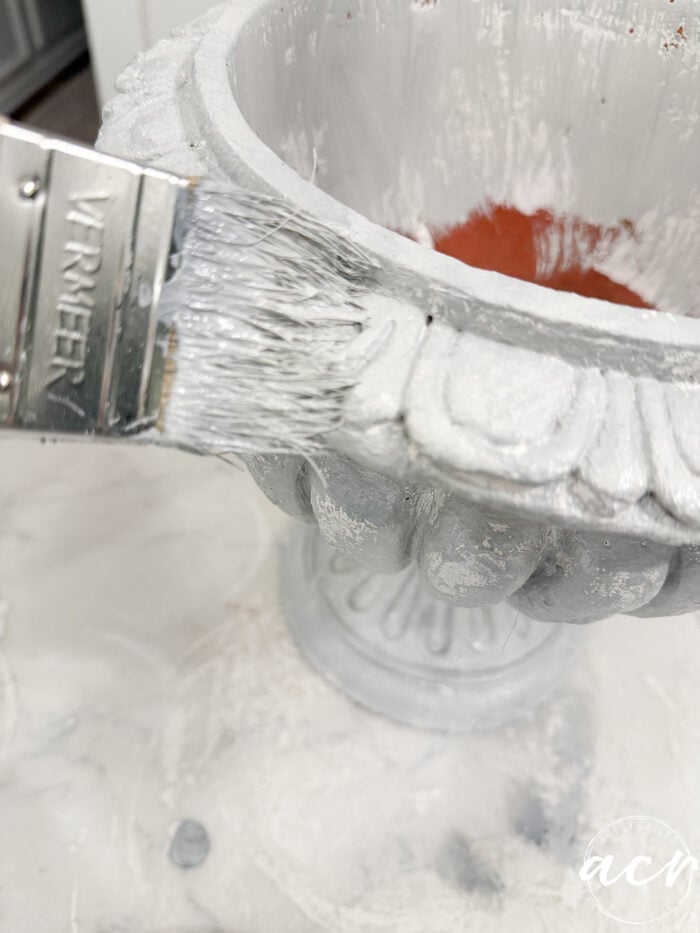 A paintbrush applying light grey faux concrete paint to a decorative terracotta urn for DIY concrete planters.