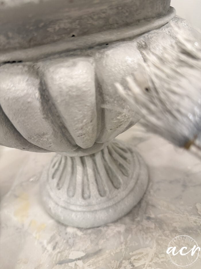 A paintbrush applying white paint to a fluted decorative urn to create a faux concrete finish.