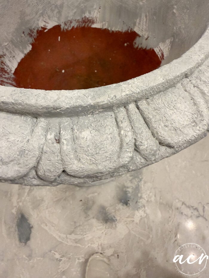 A close-up view of a large, decorative urn being painted with a thick layer of white textured paint to create DIY planters, revealing a reddish-brown interior.