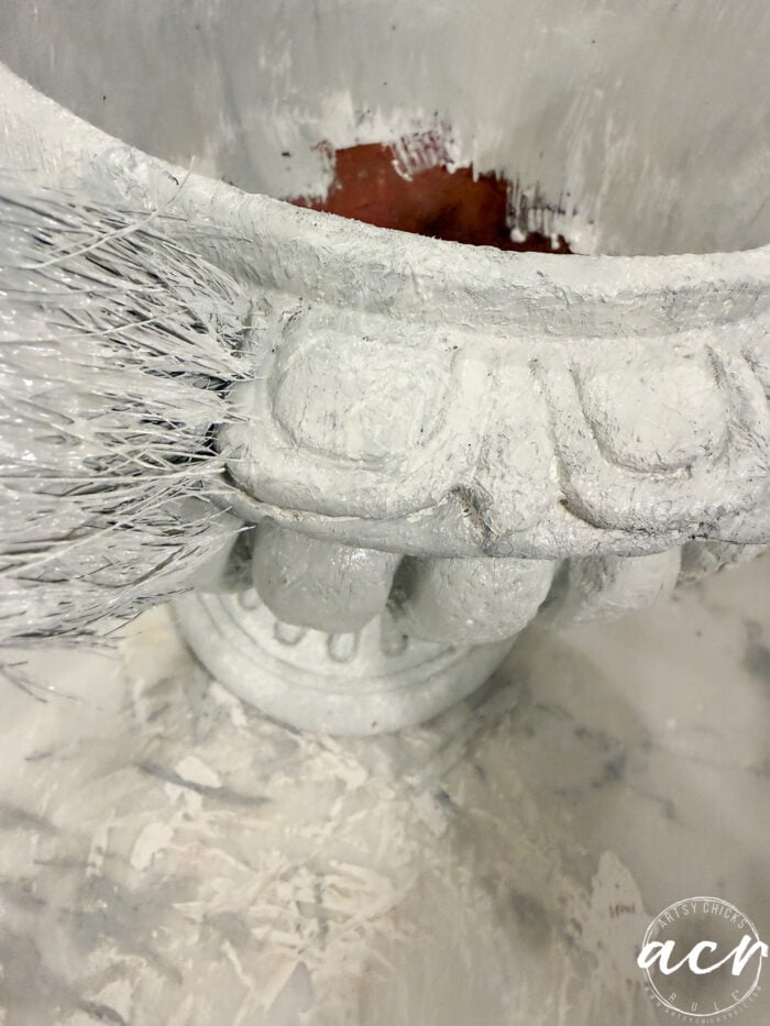 A paintbrush applying white paint to an ornate faux concrete garden urn during a DIY concrete planter tutorial.