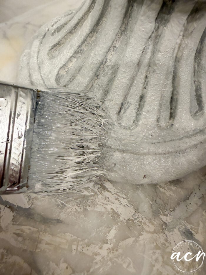 A paintbrush applying white paint to a textured, fluted object for DIY concrete planters.