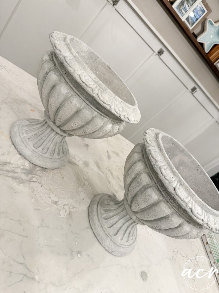 Two grey, faux concrete pedestal urns with a weathered look sitting on a white marbled surface.