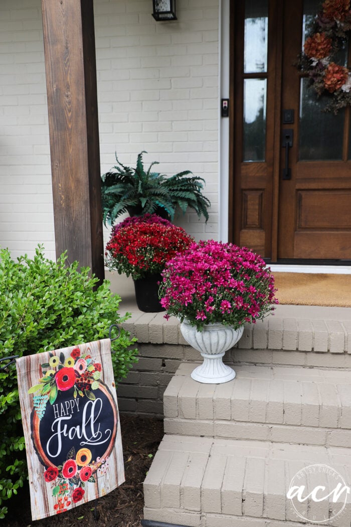 A front porch featuring a Happy Fall garden flag, potted pink and red mums in DIY concrete planters, and a wooden front door with a wreath.