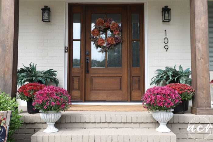 Front door with a floral wreath, flanked by pink and red chrysanthemums in DIY concrete planters on brick steps, with the house number 109 on the wall.