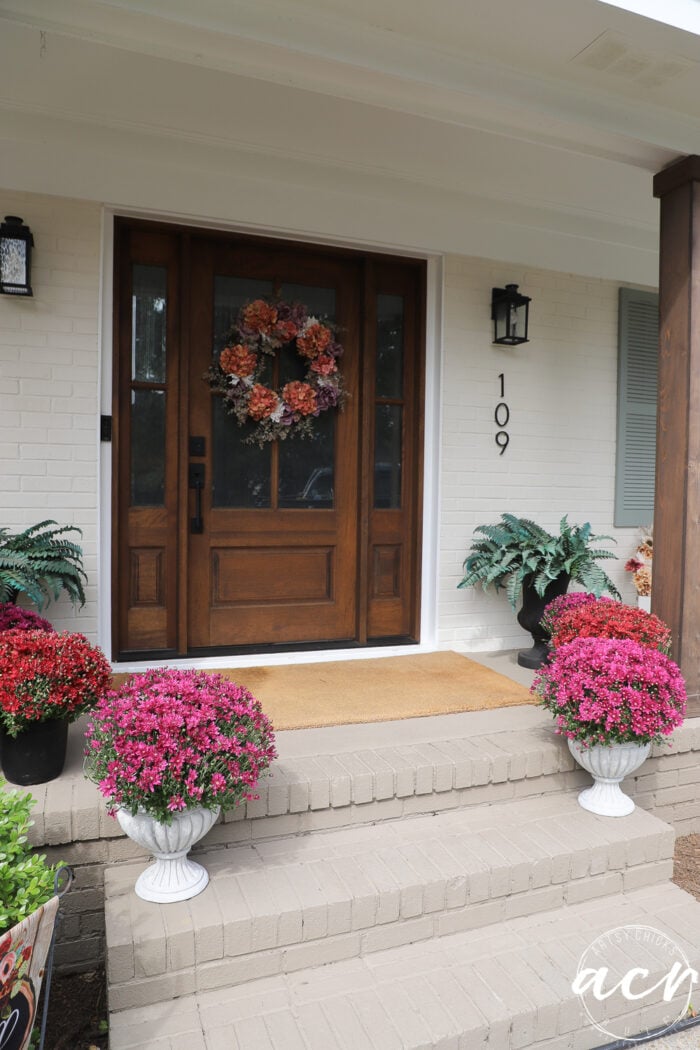 Front porch with a dark wood front door, a floral wreath, the house number 109, and several DIY concrete planters filled with pink and red chrysanthemums on the brick steps.
