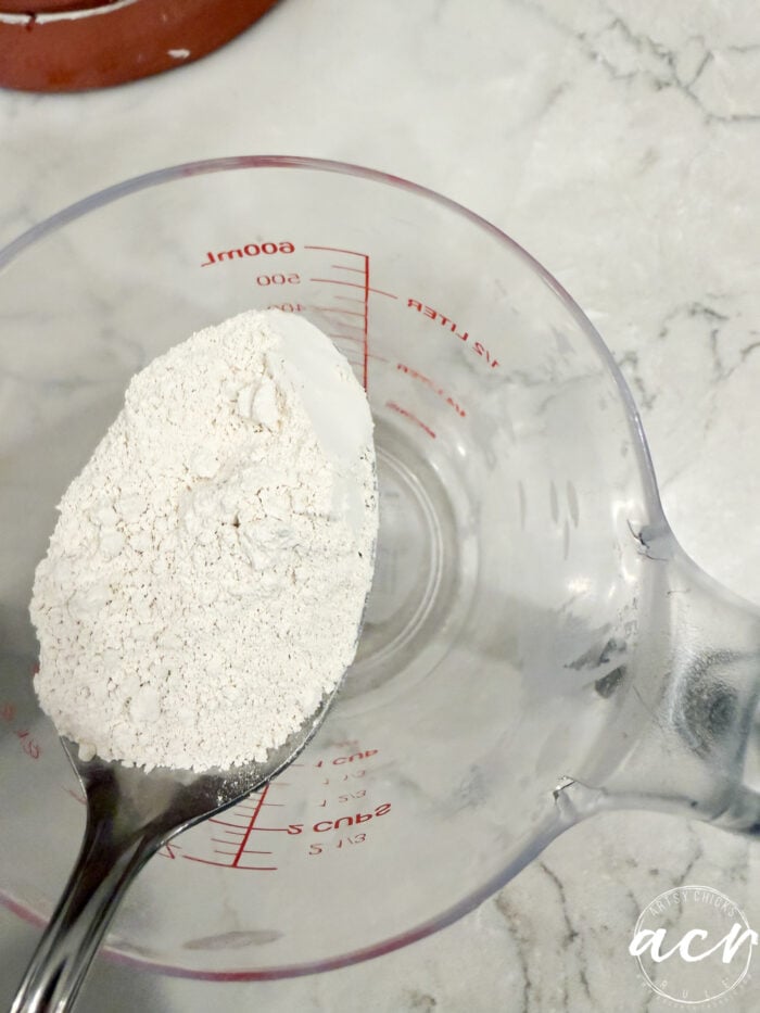 A metal spoon filled with white flour for a faux concrete project held over a plastic measuring cup.