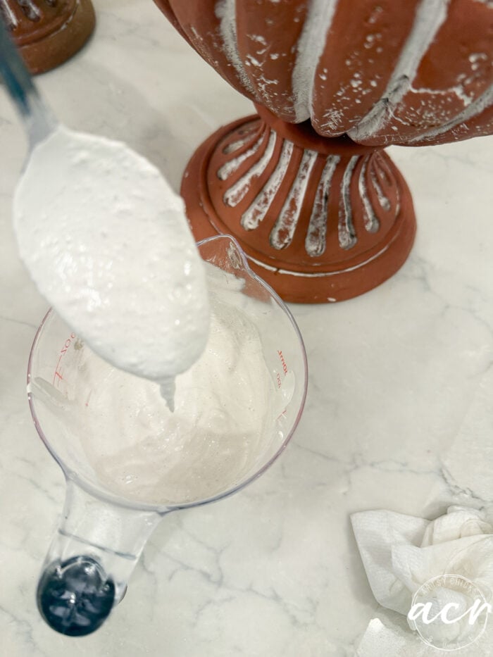 A metal spoon drips white paint into a clear measuring cup next to a weathered terracotta urn being transformed into a DIY planter.