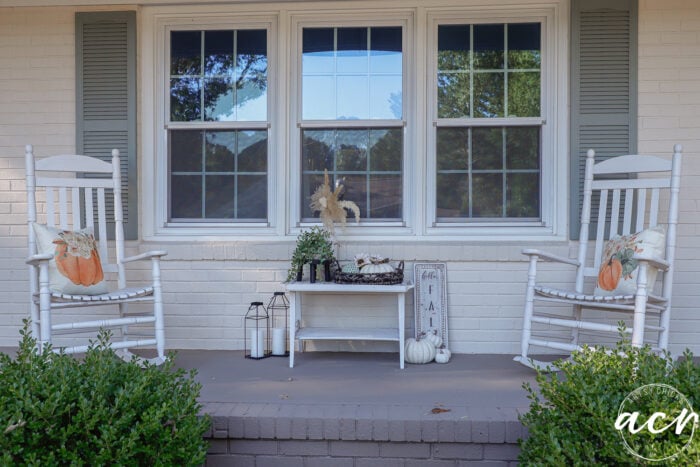Two white rocking chairs with pumpkin pillows sit on a porch on either side of a small table decorated with pumpkins, greenery, and a sign that reads Hello Fall, creating a simple fall porch.