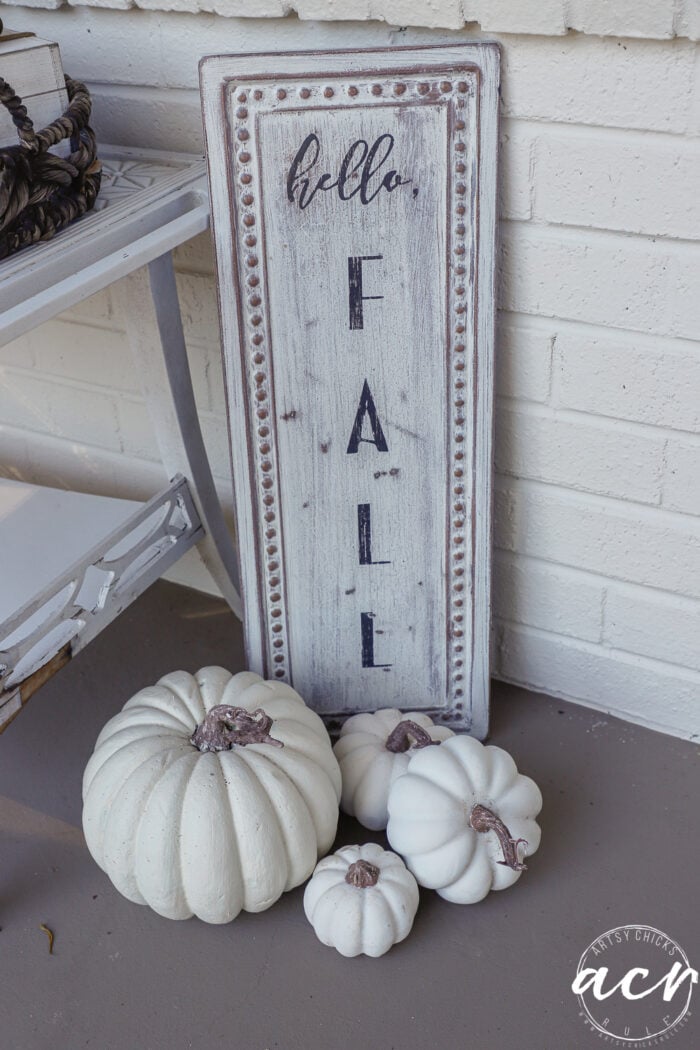 A vertical wooden sign reading hello, FALL leans against a white brick wall next to four white pumpkins, offering simple fall decor for a porch.