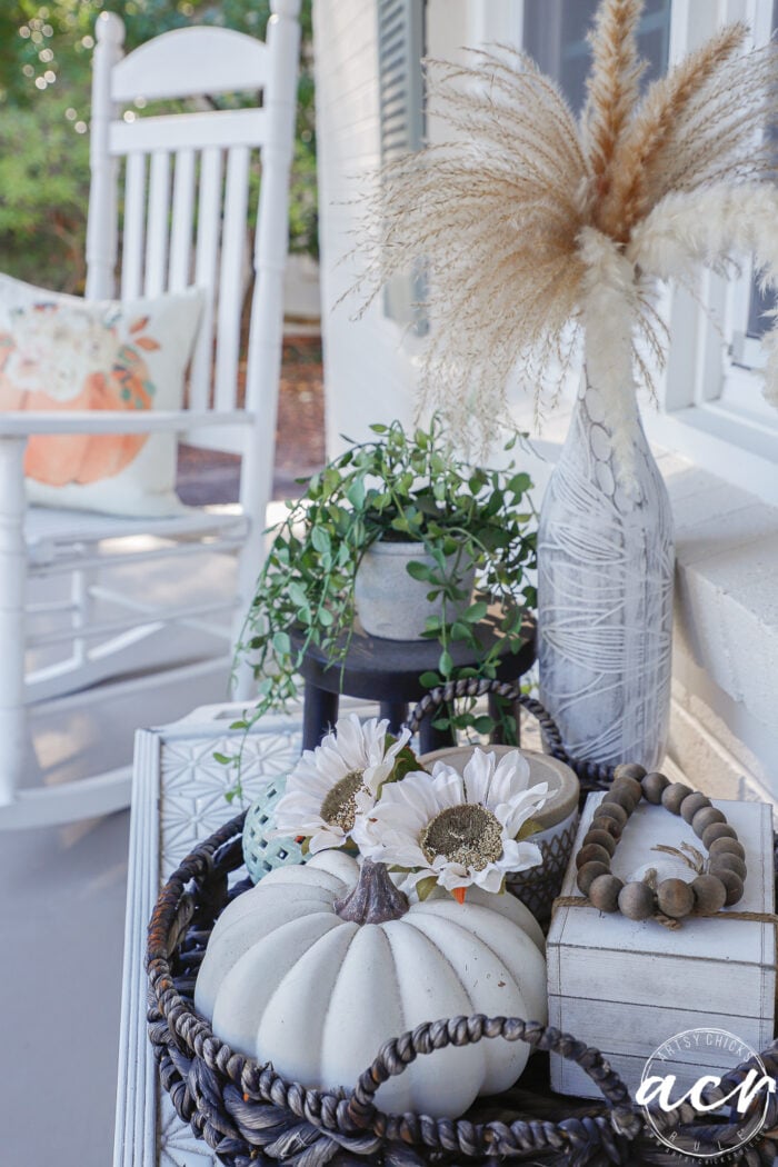 A wicker tray on a porch table holds simple fall decor, including a white pumpkin, two faux white flowers, a wooden bead garland, and a textured vase with dried pampas grass.