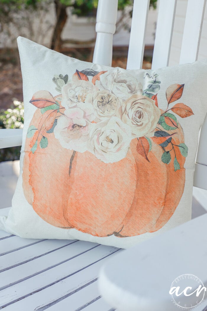 A decorative throw pillow featuring a watercolor-style orange pumpkin topped with a floral arrangement, resting on a white wooden chair as part of simple fall decor.