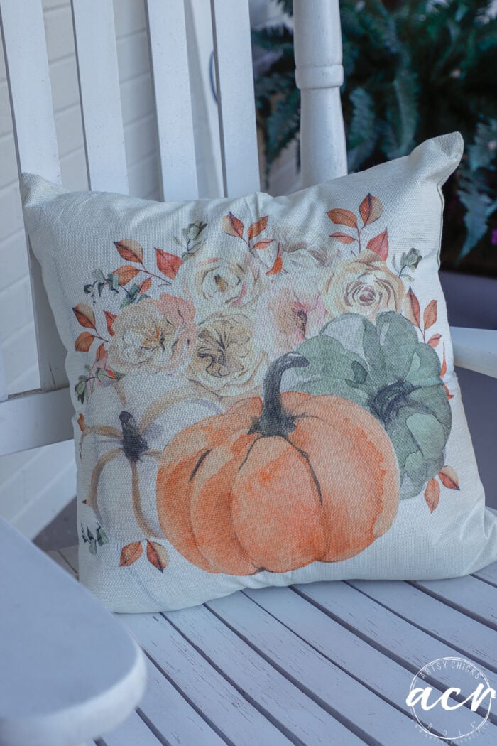 Square throw pillow on a white chair, featuring a watercolor print of an orange pumpkin, a green pumpkin, and fall-colored flowers for simple fall decor.